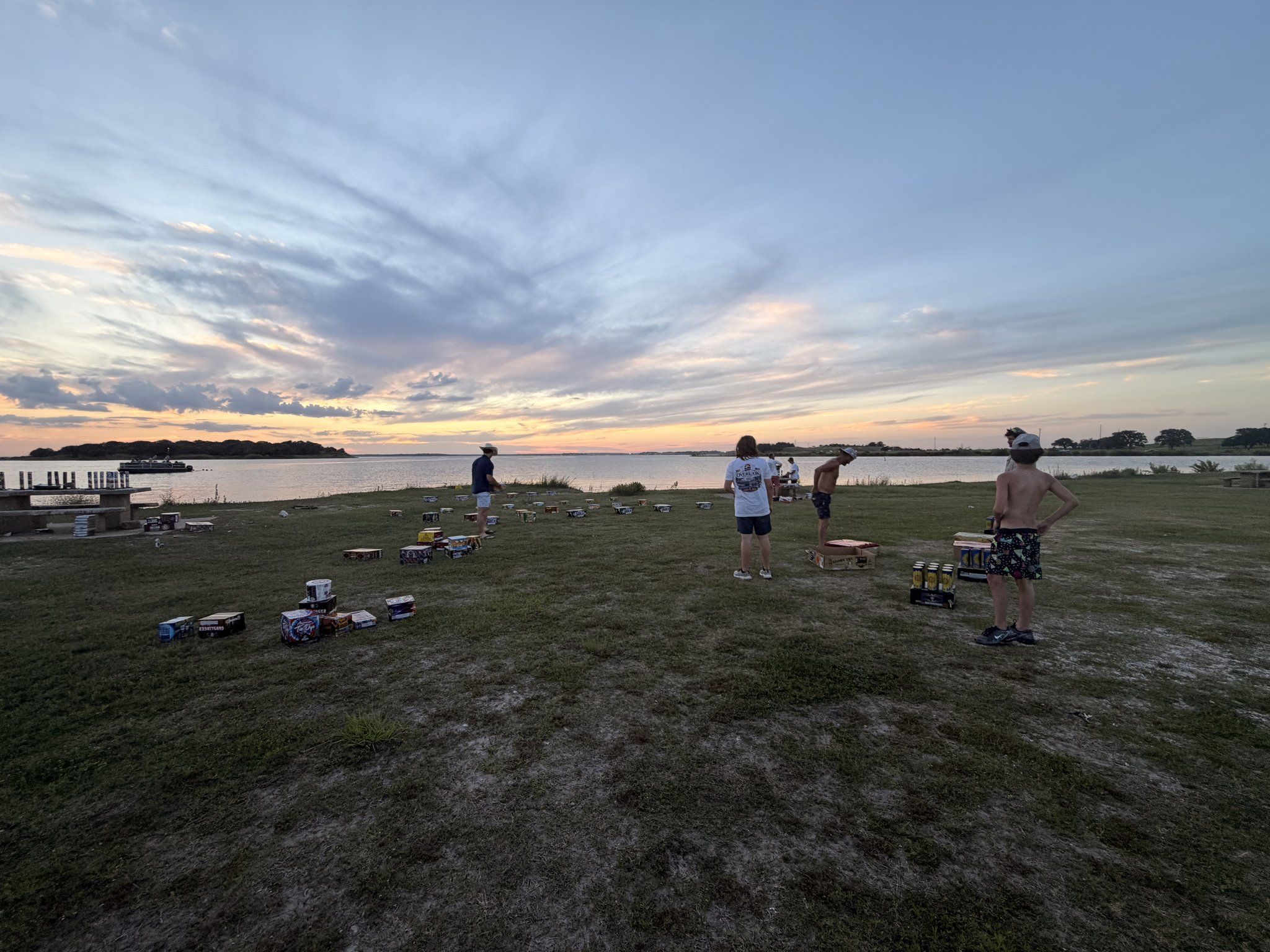 A washer-pitching tournament laid out across the lakefront lawn at dusk with players mid-game