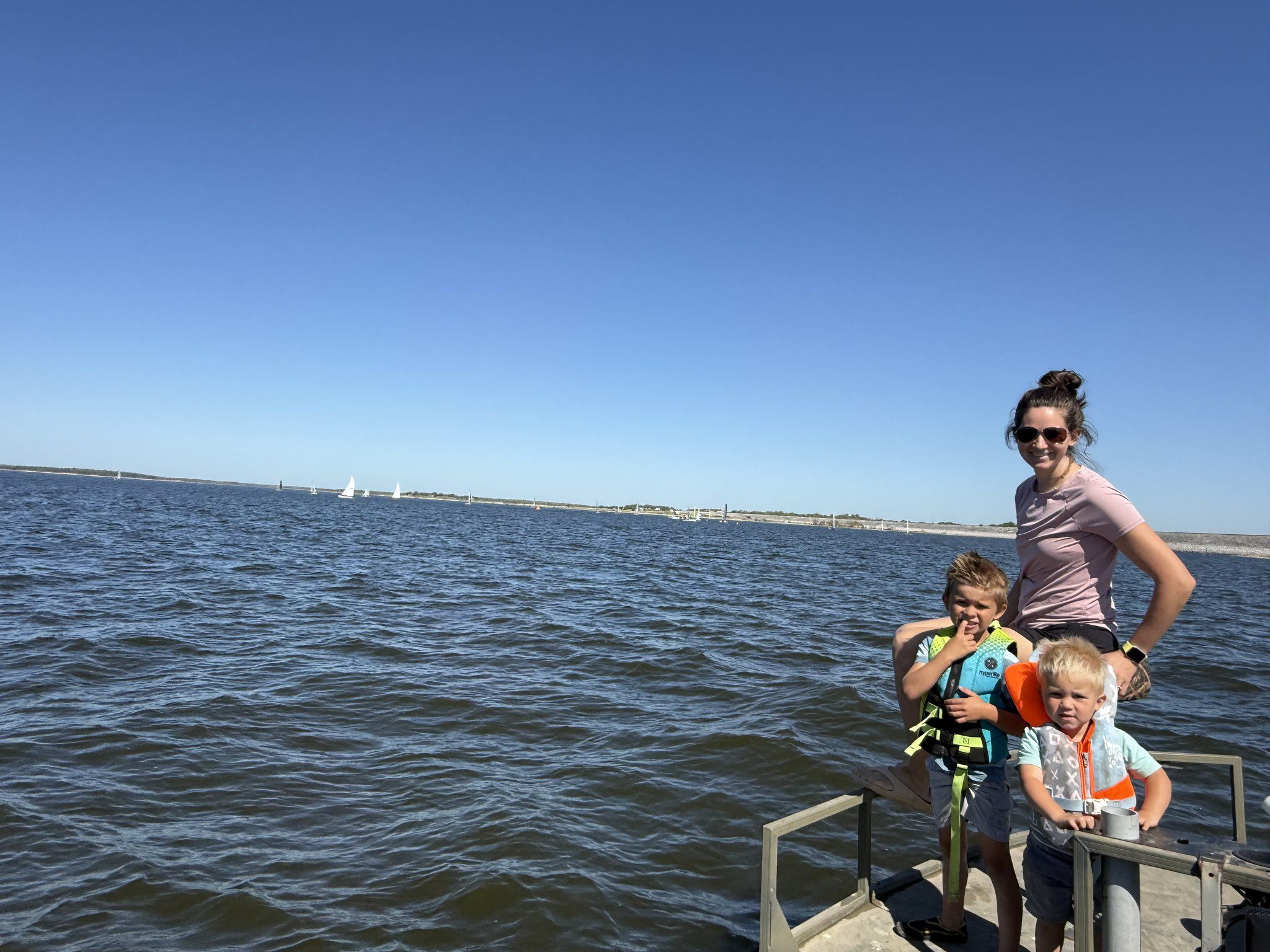 A woman on the dock with two small boys in bright life jackets, open water and distant sailboats behind