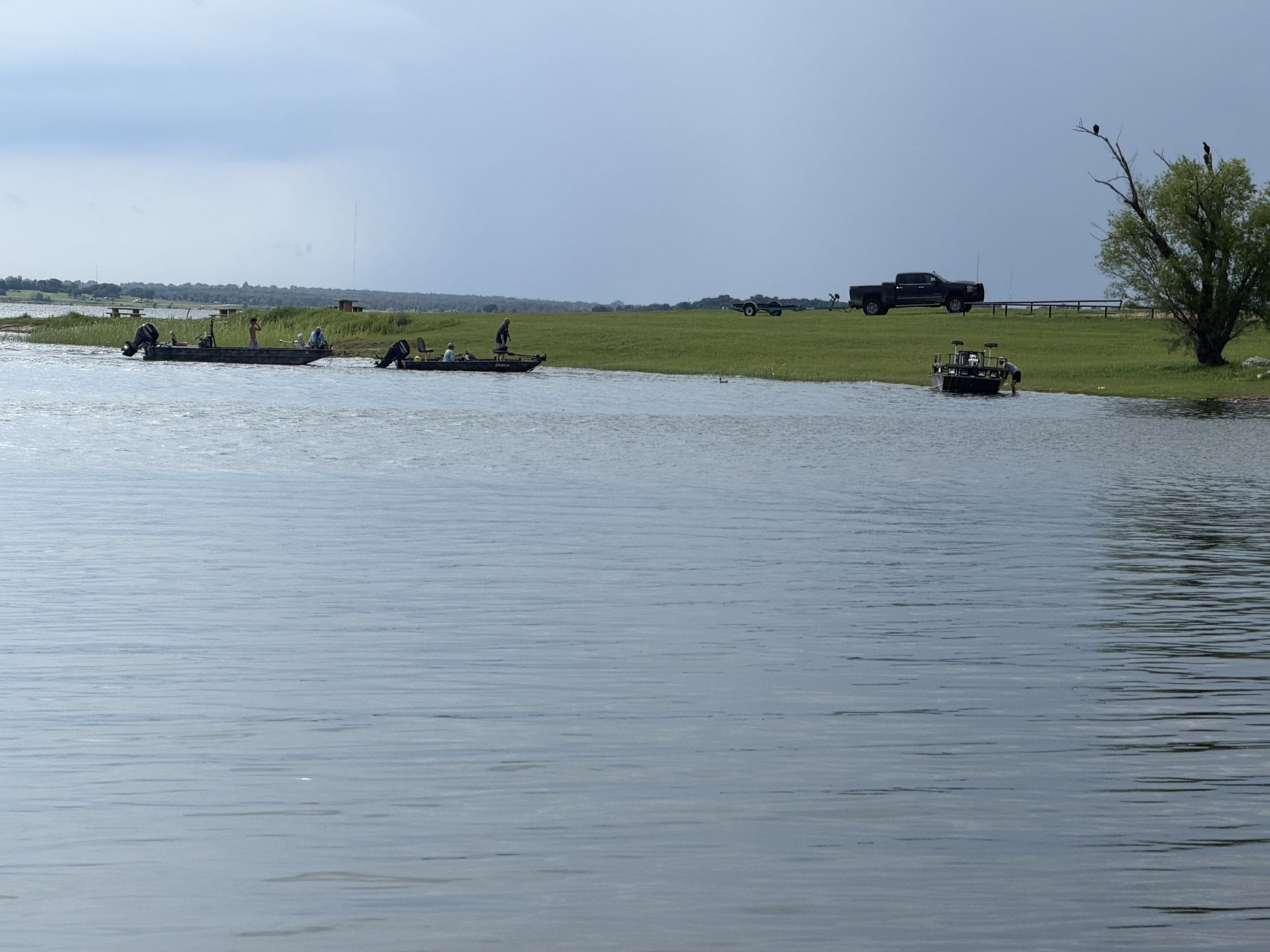 Half a dozen bass boats spread across the water near the bank with anglers aboard, trucks and trailers lined along the shore behind.