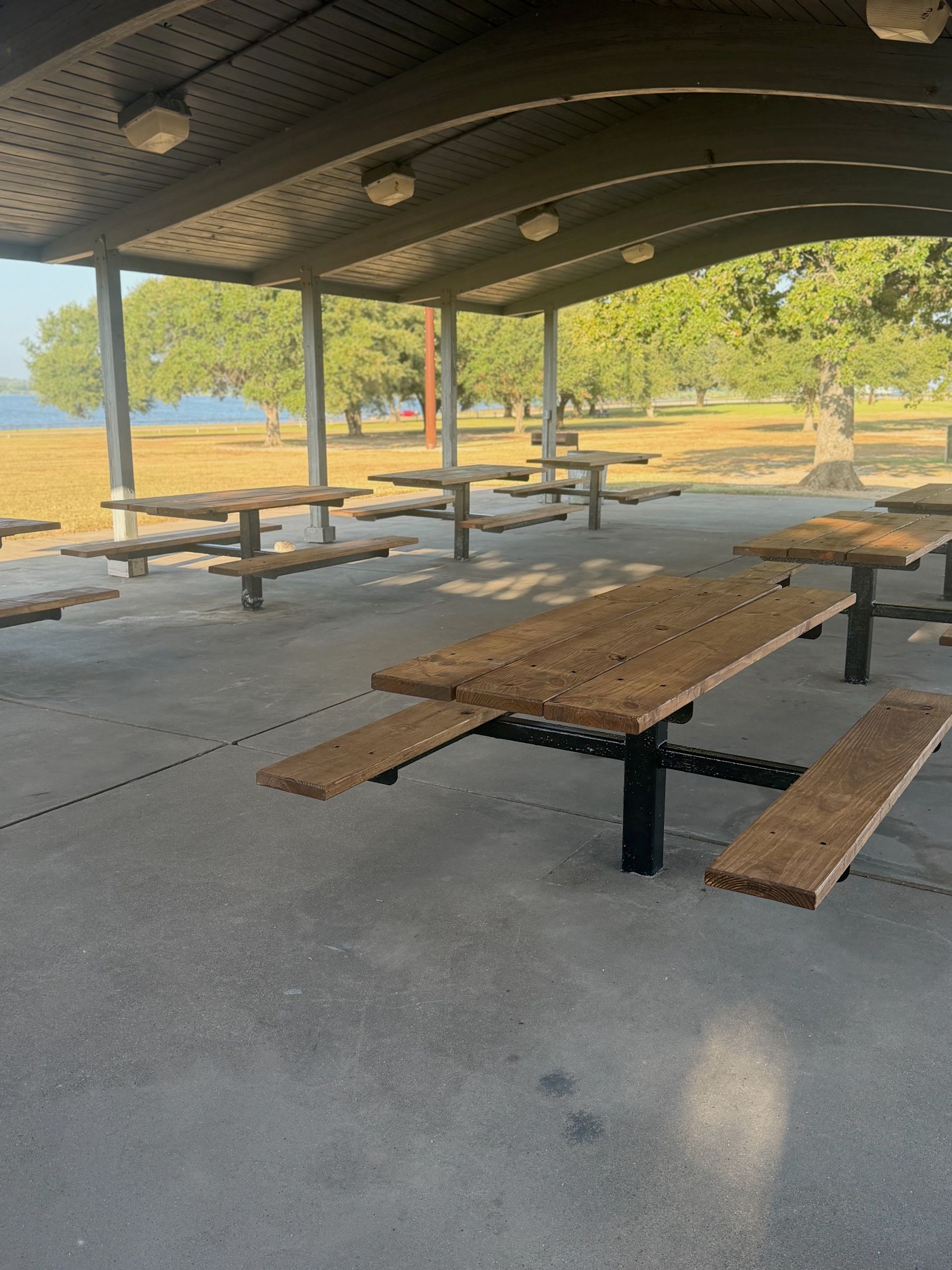 Picnic tables on the concrete floor of the pavilion, with shade oaks and the water in the distance.
