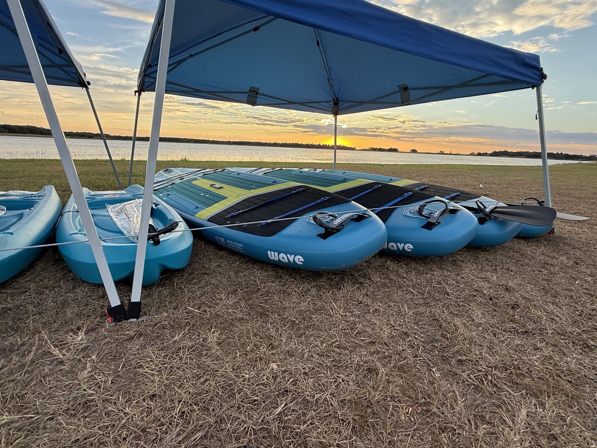 Paddleboards and kayaks stacked under a canopy with the sun setting over the water.